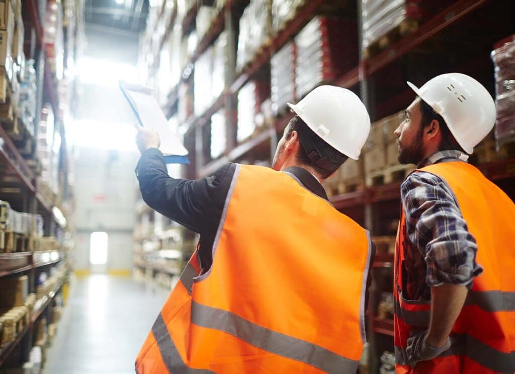 Wholesaler and Distribution Insurance - Revision Managers Discussing Inventory of Goods in a Large Warehouse While Holding a Clipboard and Wearing Safety Vests and Hard Hats